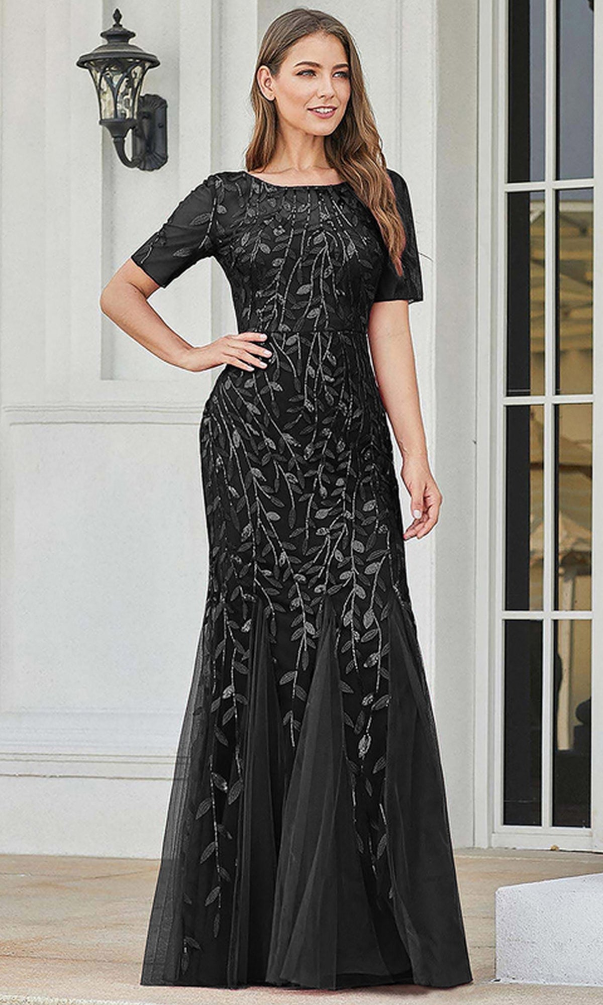 A woman stands before a white building wearing the Short-Sleeve Modest Long Mermaid Prom Dress 7707, a floor-length black formal gown with leaf-patterned embroidery and short sleeves.